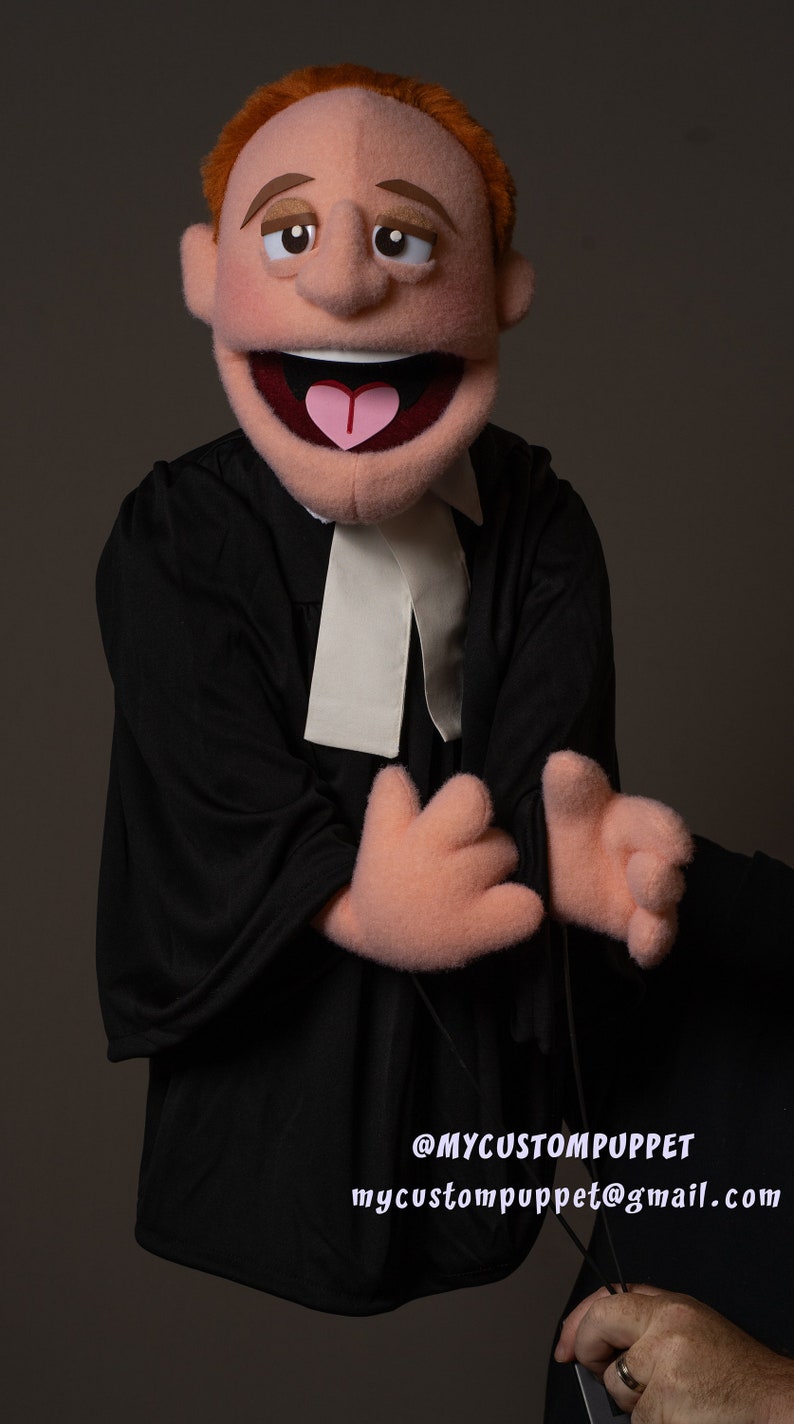May include: A red-haired puppet wearing a black robe with a white collar. The puppet has a big smile and is holding its hands up in front of it. The puppet is being held by a hand with a ring on it. The text on the image reads "@MYCUSTOMPUPPET mycustompuppet@gmail.com".