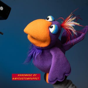 May include: A handmade purple bird puppet with an orange beak, blue and white eyes, and colorful feather accents. The puppet has a fluffy purple neck and wings. The text "HANDMADE BY @MYCUSTOMPUPPET" is displayed in a red rectangle.