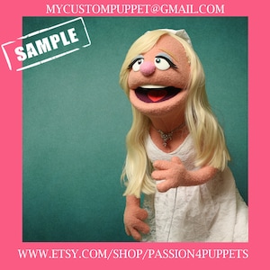 May include: A custom-made puppet with blonde hair, wearing a white dress and a necklace. The puppet has a pink face with a wide smile and blue eyes. The background is a teal green color.