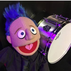 May include: A blue-haired puppet with a red mouth and a pink heart on its tongue is sitting next to a silver snare drum with a white drumhead. The drum has a black sticker with the letters "DJS" on it.