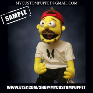May include: A handmade puppet with a yellow face, brown hair, and a red baseball cap. The puppet wears a white t-shirt with a black dog graphic and black shorts. The word "SAMPLE" is in a white box in the upper left corner. The Etsy shop URL is at the bottom.