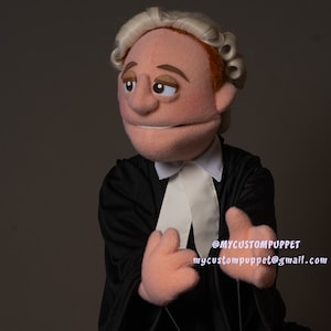 May include: A hand puppet dressed as a lawyer, wearing a black robe, white collar, and a blonde wig. The puppet has a serious expression and is looking to the left. The puppet's hands are visible and are in a gesture of speaking. The text "@MYCUSTOMPUPPET mycustompuppet@gmail.com" is visible in the bottom right corner of the image.
