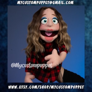 May include: A red and black plaid shirt puppet with long brown hair and blue eyes. The puppet is smiling with its tongue sticking out. The puppet is wearing a red and black plaid shirt and has a hand puppet hand.
