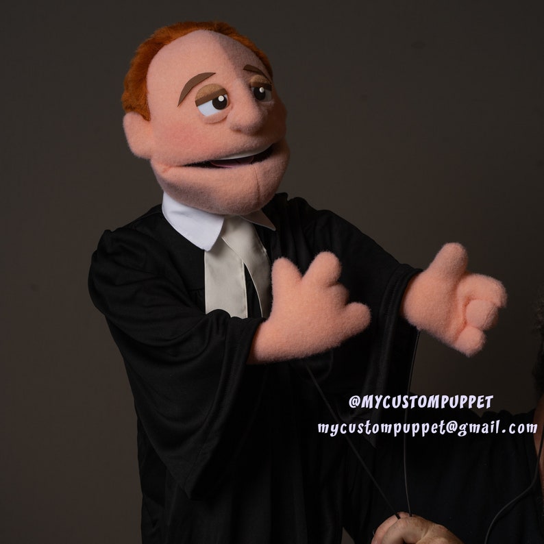 May include: A hand puppet dressed as a lawyer, wearing a black robe and white collar. The puppet has a red wig, brown eyes, and a smiling expression. The puppet's hands are outstretched, and the puppet is looking to the right. The text "@MYCUSTOMPUPPET mycustompuppet@gmail.com" is visible in the bottom right corner of the image.
