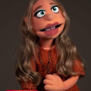 Op de afbeelding: Handgemaakte pop met een perzikkleurig gezicht, blauwe ogen en lang blond haar. De pop draagt een roestkleurige kanten top en een kralenketting. De tekst "HANDMADE BY @MYCUSTOMPUPPET" is zichtbaar.