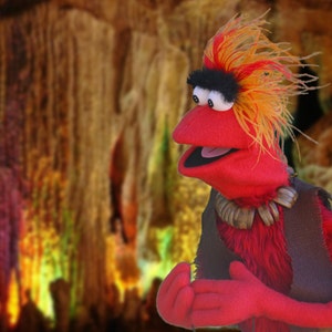 Op de afbeelding: Een rode pop met oranje en geel haar, een zwarte haarband en een ketting van witte kralen. De pop draagt een donker vest. De achtergrond is een vage grot met kleurrijke verlichting.