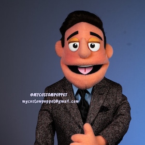 Puede incluir: Una marioneta hecha a medida de un hombre con un traje de tweed gris oscuro, una camisa azul y una corbata negra. La marioneta tiene una cara color melocotón, cabello castaño y ojos grandes y expresivos. La boca de la marioneta está abierta, revelando una lengua rosada. El texto "@MYCUSTOMPUPPET" y una dirección de correo electrónico son visibles.
