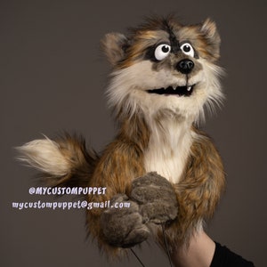 Könnte beinhalten: Eine Handpuppe eines Waschbären mit braunem, beigem und weißem Fell. Die Puppe hat große, ausdrucksstarke Augen, eine schwarze Nase und einen lächelnden Mund. Die Pfoten der Puppe sind grau, und der Text "@MYCUSTOMPUPPET mycustompuppet@gmail.com" ist sichtbar.
