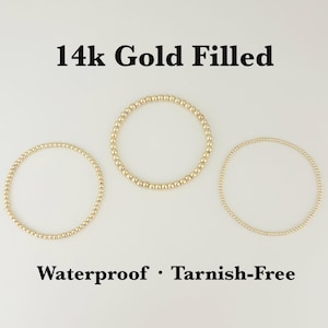 以下が含まれることがあります： 白い背景に、3つのゴールドフィルドビーズブレスレットが表示されています。 ブレスレットはビーズのサイズが異なり、上部と下部に「14k Gold Filled」と「Waterproof • Tarnish-Free」の文字があります。
