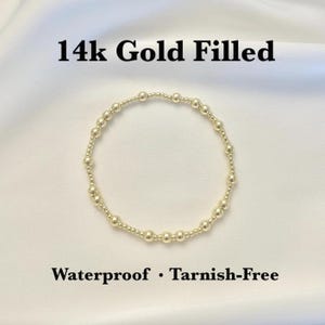 Może przedstawiać: Złota bransoletka z koralików. Bransoletka składa się z małych, okrągłych koralików nanizanych na sznurek. Tekst "14k Gold Filled" i "Waterproof • Tarnish-Free" jest wyświetlany nad i pod bransoletką.