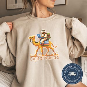 Könnte beinhalten: Beigefarbener Pullover mit einer pixelartigen Illustration einer Person, die auf einem Kamel mit einem Laptop reitet. Der Text "Digital Nomad" ist unter dem Bild gedruckt.