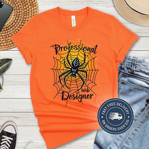 May include: Bright orange t-shirt featuring a black spider web design with a black spider in the centre. The text "Professional Web Designer" is above and below the spider. The shirt is folded on a surface with a pair of jeans.