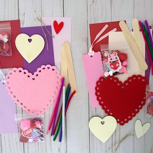 May include: A collection of Valentine's Day craft supplies, including pink and red heart-shaped felt cutouts, wooden hearts, pipe cleaners, popsicle sticks, and small bags of embellishments. The items are arranged on a light wood-grain surface.