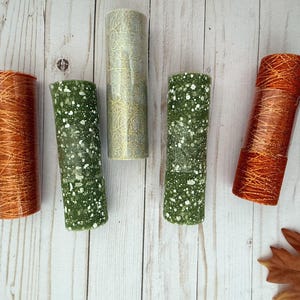 May include: Five cylindrical decorative candles in shades of green, gold, and orange. The candles have textured surfaces with speckled and patterned designs. Autumn leaves are scattered around the candles on a light wood surface.