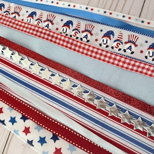 May include: Assortment of patriotic ribbons in red, white, and blue. Designs include stars, stripes, gingham, and gnome illustrations with "USA" text. Some ribbons have silver star embellishments.