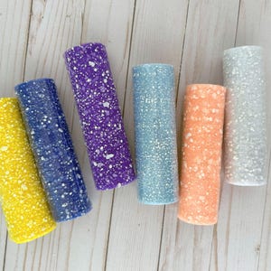 May include: An overhead shot displays six cylindrical objects in various colors: yellow, navy blue, purple, light blue, coral, and silver. Each cylinder is speckled with white dots, creating a textured appearance. The objects are arranged on a light-colored wooden surface.