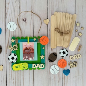 May include: A green and blue picture frame with a photo of two hands forming a heart. The frame is decorated with wooden sports balls, stars, and the words "I Love You" and "Dad".