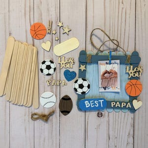May include: A DIY craft kit featuring a blue popsicle stick frame with a twine hanger, a photo clip, and wooden cutouts including basketballs, soccer balls, a football, stars, hearts, and the text "BEST PAPA" and "I love you".