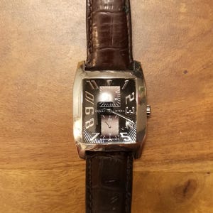 May include: A rectangular wristwatch with a silver-tone stainless steel case and a dark brown embossed leather strap. The black dial features large white numerals 8, 9, 10, 2, 3, 4, 5, 6, 7, the text GUESS STEEL, and two sub-dials with silver hands.