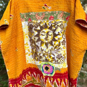 May include: A mustard yellow quilted top featuring a central white panel with a brown sun graphic. The garment includes a green floral border at the top, a red patterned hem at the bottom, and a small circular floral applique detail on the front.