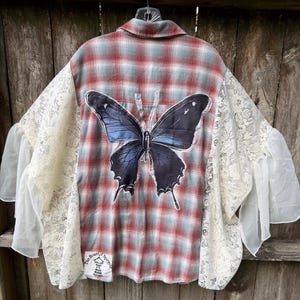May include: A button-down shirt featuring a red and grey plaid pattern with a large black and blue butterfly graphic on the back. The garment includes cream-colored lace and sheer fabric panels on the sleeves, and a small white label at the bottom.