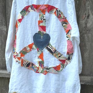 May include: A white long-sleeved shirt featuring a large peace sign applique on the back, constructed from various colorful patterned fabric scraps. A solid dark blue denim heart is centered within the peace sign. A small label reads The House of Upcycle.