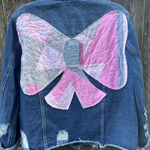 May include: A distressed denim jacket with a large, quilted bow design on the back. The bow features pink and gray patchwork, adding a unique, handmade touch. The jacket has a classic collar, button closures, and distressed details.