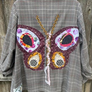 May include: A brown plaid blazer with a large butterfly applique on the back. The butterfly is made of various patterned fabrics in shades of purple, yellow, and white, with black and gold accents. The blazer has long sleeves.