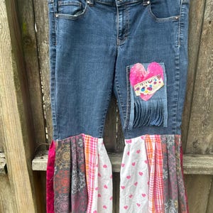 May include: Blue denim jeans with patchwork heart and fringe detail. The jeans feature flared bottoms with patterned fabric in pink, white, and gray. A pink heart patch is sewn on the front, adding a unique, handcrafted touch.