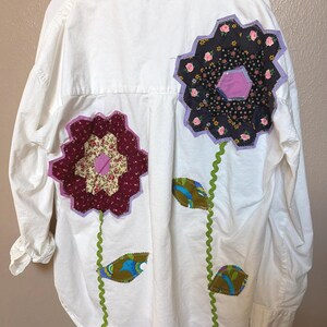 May include: White button-down shirt with floral patchwork design on the back. The flowers are made of various fabrics, including floral prints and solid colors. Green stems and leaves add to the design. The shirt has long sleeves and a classic collar.