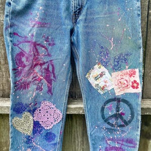 May include: Blue denim jeans with painted purple floral designs, paint splatters, and patches. The jeans feature a peace sign and the word "LOVE" painted on the leg. Patches include floral and patterned fabric.
