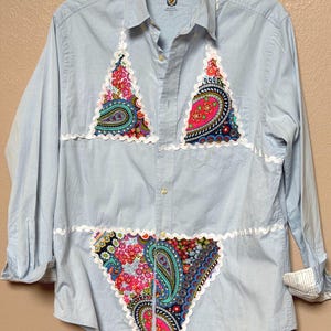 May include: A light blue long-sleeve button-down shirt featuring a Club Room label at the collar. The front is decorated with three triangular fabric patches in a colorful paisley and floral pattern, each outlined with white rickrack trim.
