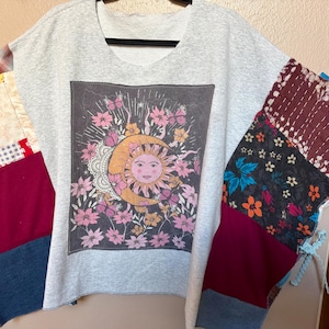 May include: A light gray short-sleeve top featuring a central graphic of a sun and crescent moon surrounded by pink flowers and butterflies. The sleeves are constructed from a patchwork of various fabrics including plaid, floral, and solid maroon panels.