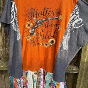 May include: Orange t-shirt with the text "Hotter than a Pistol" and a gun graphic, surrounded by flowers. The sleeves are gray with white feather and floral designs. The bottom hem features colorful fabric strips.