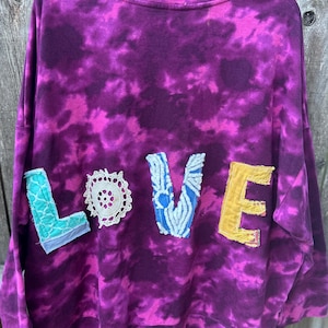 May include: A purple tie-dye sweatshirt with the word "LOVE" appliqued across the front. The letters are made from various fabrics, including a teal patch, a white doily, a blue patterned fabric, and a yellow patch. The sweatshirt is hanging on a black hanger.
