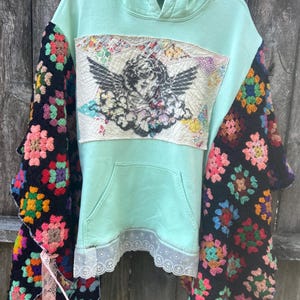 May include: A mint green hoodie with a front pocket and a hood. The sleeves are made of black crochet with colorful floral patterns. A patch with an angel illustration is sewn on the front, and the bottom hem has a white lace trim.