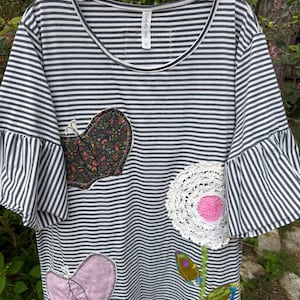 May include: A grey and white striped short-sleeve shirt with ruffled sleeves and a round neckline. The shirt features three fabric appliqués: a dark floral butterfly, a pink butterfly, and a white crocheted flower with a pink center.