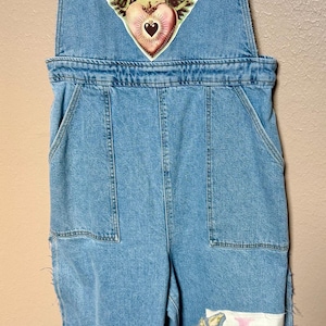 May include: Light blue denim overalls featuring a graphic of a winged figure with a crown and heart on the chest. The legs include a pink heart patch, floral appliqués, and a white rectangular patch with the vertical pink text LOVE.