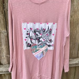 May include: A long-sleeved, dusty rose-colored shirt with a crew neck. The shirt features a white patch with a Cupid graphic, pink hearts, and a fabric heart applique. The shirt has a relaxed fit and a casual style.
