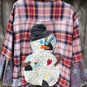 May include: A pink, red, and blue plaid flannel shirt with a quilted snowman applique on the back. The snowman has a black hat, blue eyes, an orange nose, and a red heart. The sleeves have patterned fabric cuffs.