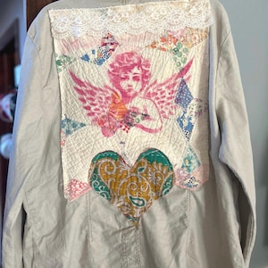 May include: A light beige jacket with a patchwork design on the back. The patchwork features a pink cherub, colorful fabric pieces, a white lace trim, and a heart-shaped design with paisley patterns.