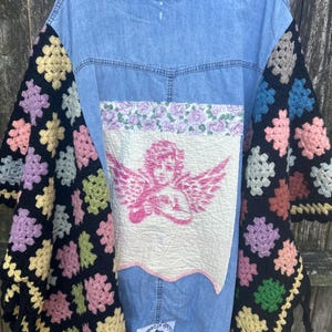 May include: A light blue denim jacket with crochet sleeves in various pastel colors. The back features a patch with a pink cherub design and floral trim. The jacket has a collar and button-up front.