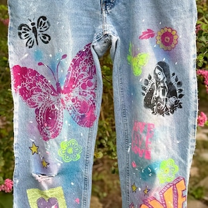 May include: Light blue denim jeans featuring colorful screen-printed designs including a large pink and purple butterfly, a black and white portrait of the Virgin Mary, the word LOVE in orange and pink, and various floral, star, and heart motifs.