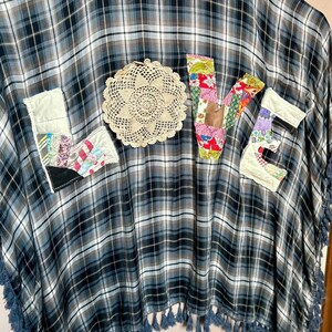 May include: A plaid poncho in shades of blue, gray, and white, with a fringed hem. The word "LOVE" is appliquéd across the front using fabric patches and a crocheted doily. The patches feature floral and patterned fabrics.