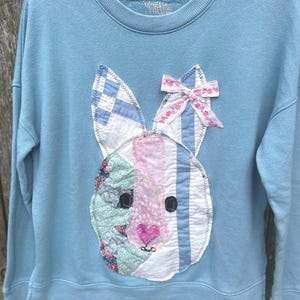 May include: Light blue sweatshirt featuring a patchwork bunny face. The bunny has striped ears, a pink nose, and a floral bow. The design is stitched onto the front of the sweatshirt, creating a unique, handmade look.