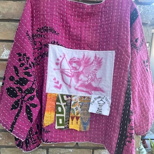 May include: A magenta-colored, quilted jacket with black floral patterns on the sleeves. A white patch features a pink Cupid design. The jacket has a patchwork of colorful fabric strips.