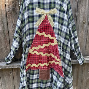 May include: A long-sleeved plaid shirt dress in green, black, and white, with a red and gold Christmas tree applique on the back. The tree has gold wavy trim and a brown square base, topped with a gold bow.