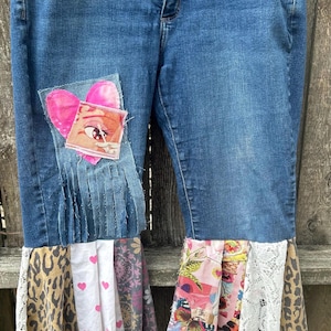 May include: Blue denim jeans with patchwork details. Features a pink heart and eye patch, and a flared hem with leopard print, floral, and lace panels. A unique, artistic design.