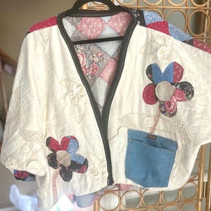 May include: Cream-colored quilted jacket with floral appliqués and a denim pocket. The jacket features a black trim and a patchwork design on the inside lining. The floral appliqués are made of various patterned fabrics in red, blue, and black.