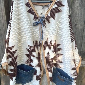 May include: Cream-colored poncho with brown geometric patterns and denim pockets. The poncho has a textured fabric, fringed hem, and tan trim. A blue ribbon ties the front closed. The poncho is hanging on a black hanger.